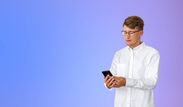 Portrait of concentrated young European businessman in white shirt looking at smartphone screen standing over purple background. Concept of social media and communication. Copy space - Fotoğraf, Görsel