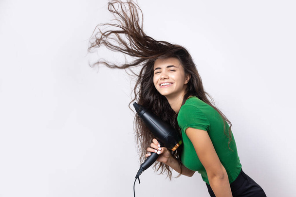 A cute young girl dressed in a green top dries her beautiful long silky hair with a hair dryer against a white background. - Foto, imagen