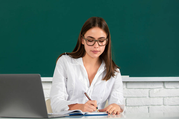Portrait of a beautiful young woman student with laptop in a university classroom. Beauty female student studying. College student on blackboard. Pretty girl student on desk. Students day - Photo, Image