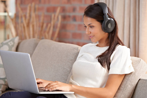 Headphones, home and a woman typing on laptop on home sofa listening to music or streaming movies. Calm female person relax on couch to listen to radio or learning a language with internet connection. - Foto, Imagen