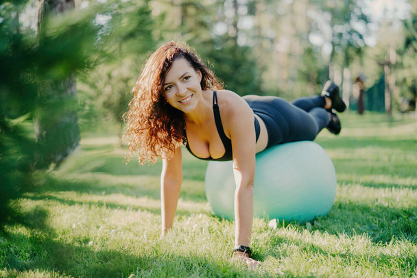 Desportista feliz no desgaste ativo usa bola de ginástica para fitness ao ar livre, posando na grama verde com um sorriso. - Foto, Imagem