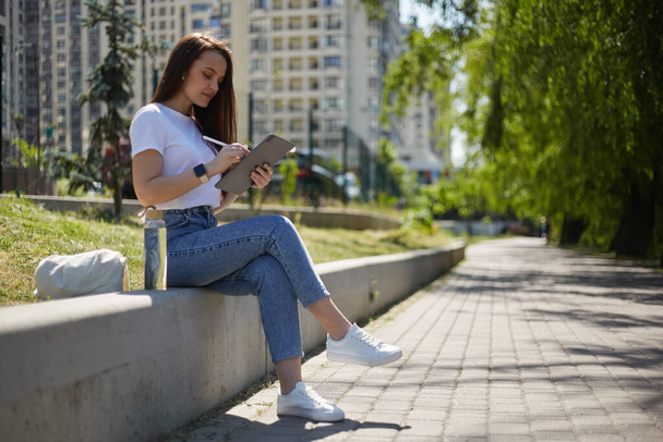 Mladý grafik sedící venku s tabletovým počítačem a kreslící skicu s digitálním perem. Ilustrátorka žena dělat náčrtek s stylus venkovní - Fotografie, Obrázek