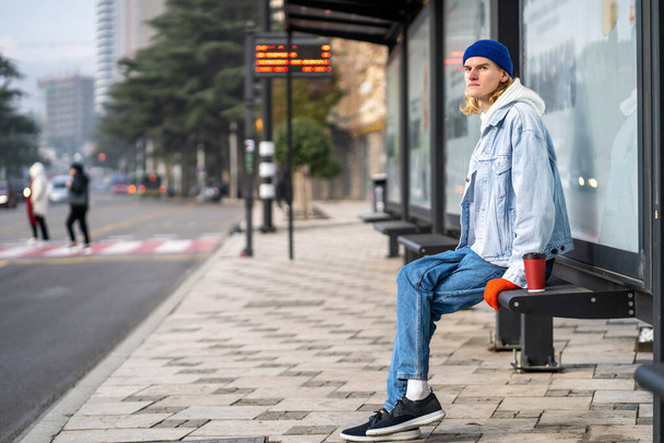 Young blond guy student waiting transport very long time in morning.Man on bus stop with coffee away. Blonde hair millennial man on bus stop in city. Modern urban transport for everyone - Foto, afbeelding