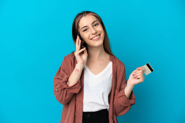 Young caucasian woman isolated on blue background keeping a conversation with the mobile phone and holding a credit card - Fotó, kép