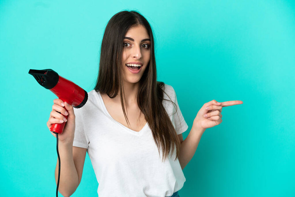 Young caucasian woman holding a hairdryer isolated on blue background surprised and pointing finger to the side - Photo, Image