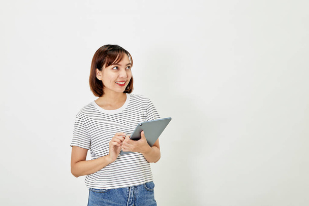 Charming young Asian female employee with short brown hair wearing t-shirt and jeans smiling and holding digital tablet standing on white background copy space - Photo, Image