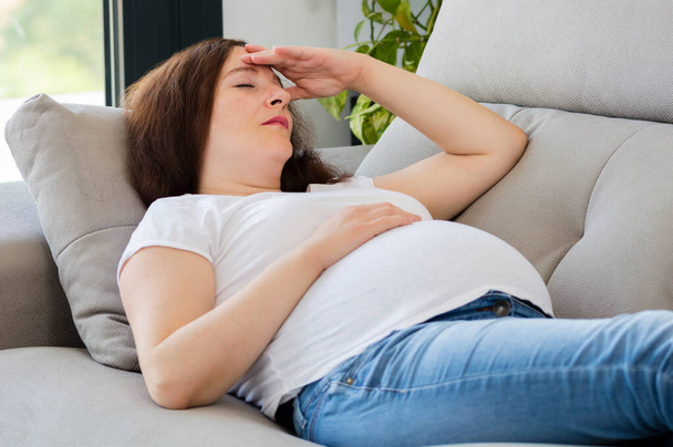 Pregnant woman with hand on forehead suffering headache lying on a couch in the living room at home - Фото, изображение
