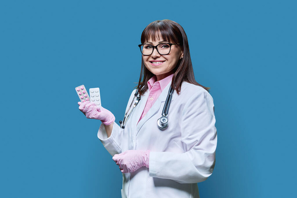 Woman doctor showing pills in blister on blue background. Friendly woman in white coat with medical gloves looking at camera. Pharmacology pharmaceutics medicine medication professional recommendation - Foto, immagini
