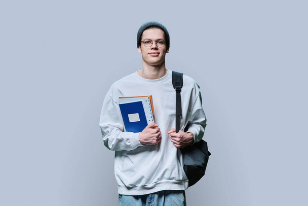 Young guy college student with backpack holding notebooks textbooks, in knitted hat eyeglasses white sweatshirt looking at camera on grey studio background. Education, studying, learning concept - Foto, Imagen