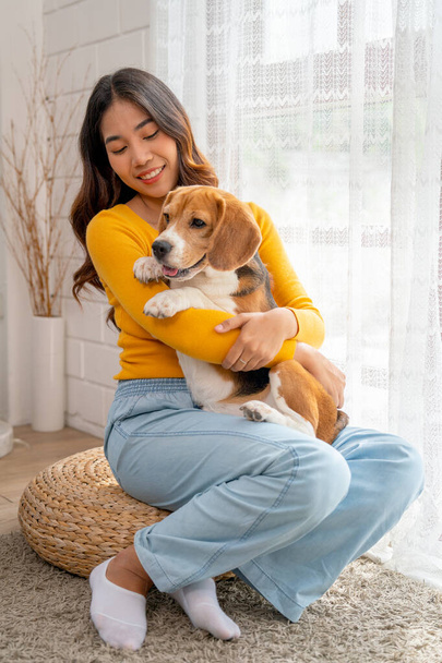 Κάθετη εικόνα της νεαρής Ασιάτισσας κορίτσι κρατήσει και να αγκαλιάσει το σκυλί beagle και να καθίσει μπροστά από την γυάλινη πόρτα στο σπίτι της και φαίνεται ευτυχής να παίξει διασκέδαση μαζί. - Φωτογραφία, εικόνα