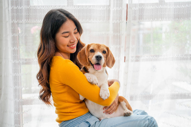 Закрийте молоду азіатську дівчину, обійміть собаку - бігера і сидіть перед скляними дверима в її будинку, і вона виглядає щасливою бавитися разом.. - Фото, зображення