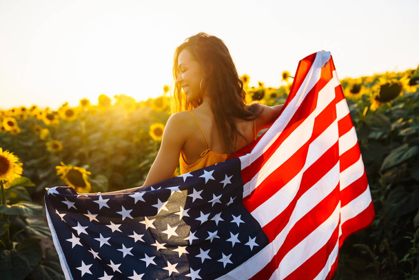 Beautiful woman holding an American flag in a sunflower field at sunset. Patriotic holiday. Independence Day, July 4th. - Фото, изображение