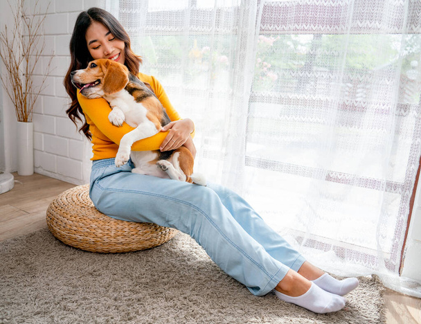 Fiatal ázsiai lány tart és ölel beagle kutya és ül előtt üveg ajtó a házában, és úgy néz ki, boldog játszani szórakoztató együtt. - Fotó, kép