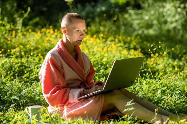 A bald woman in a bathrobe works with a laptop in nature. summer time, sunny day. - Valokuva, kuva