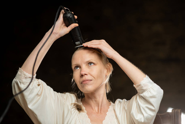 A thin middle-aged woman cuts off all her hair on her head with a clipper. dark background. - Foto, Imagem