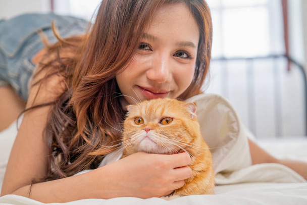 Close up beautiful Asian woman lie near her orange cat also scratch the chin of cat to make it relax and she also look at camera with smiling. - Foto, Imagen