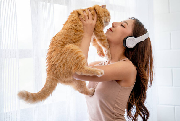 Krásná asijské dívka nosit sluchátka a držet oranžovou kočku před skleněnými okny s bílou záclonou a vypadají štěstí spolu v jejich domě. - Fotografie, Obrázek