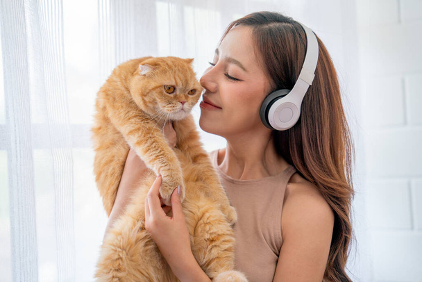 Mooi aziatisch meisje dragen oortelefoon en vasthouden en kussen kat in de voorkant van glas ramen met witte gordijn en ze kijken geluk samen in hun huis. - Foto, afbeelding
