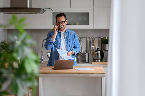 Cheerful young businessman holding document and talking over smart phone while standing in kitchen - Φωτογραφία, εικόνα