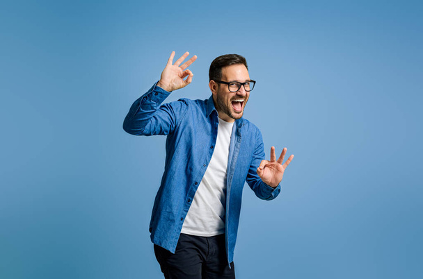 Satisfied businessman gesturing OK sign and shouting while standing against blue background - Foto, Imagen