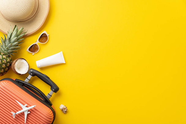 Dreams of sunny vacation. Top view arrangement with suitcase, miniature plane, beach essentials, glasses, cap, sunscreen, and luscious ananas on vivid yellow background, offering space for text or ad - Photo, Image