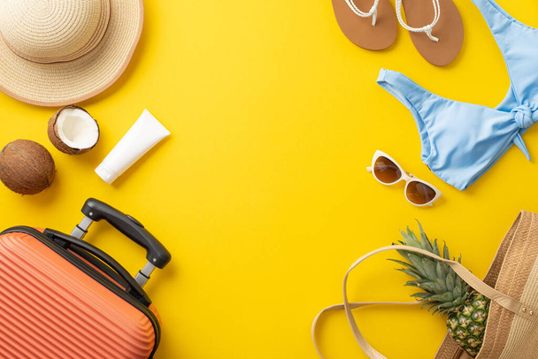 Tropical getaway theme. Overhead view of suitcase, beach essentials, bag, glasses, hat, sunscreen, flip-flops, swimwear and juicy fruits on a vibrant yellow backdrop. Perfect for text or advertising - Photo, Image