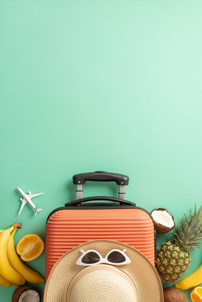 Тропическая концепция путешествия. Над вертикальным видом фото соломенной шляпы с солнцезащитными очками на оранжевом чемодане в окружении тропических фруктов и модели самолета на изолированном бирюзовом фоне с копирайтом - Фото, изображение