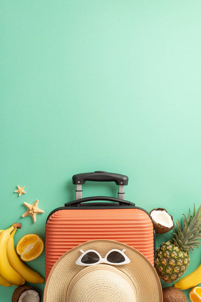 Tropical journey concept. Above vertical view photo of straw hat with sunglasses on orange suitcase surrounded by tropical fruit and starfish on isolated turquoise background with copyspace - Photo, Image