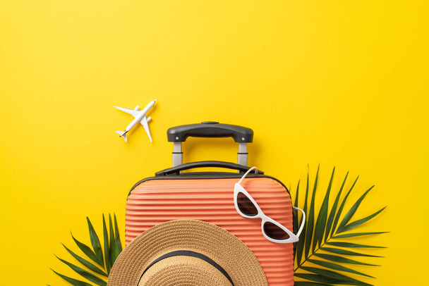 Καλοκαιρινές διακοπές. Πάνω άποψη φωτογραφία πορτοκαλί βαλίτσα με καπέλο ηλίου και γυαλιά ηλίου σε αυτό με φύλλα φοίνικα και το μοντέλο αεροπλάνο σε απομονωμένο φωτεινό κίτρινο φόντο με copyspace - Φωτογραφία, εικόνα