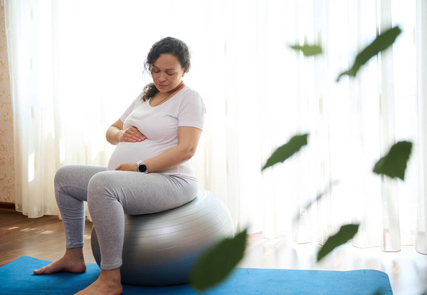 Beautiful pregnant woman, sitting on fitball, holding hands on her belly, exercising in late pregnancy at home, preparing her body for easy childbirth. Sport. Fitness. Active healthy lifestyle concept - Fotoğraf, Görsel