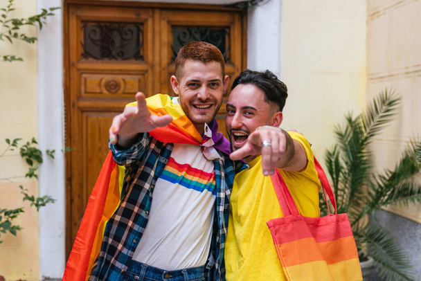 Két meleg barát, akik boldogságot és büszkeséget árasztanak, szorosan ölelkeznek, vidáman mutogatnak a díszített ajtójuk előtt, LMBT-szimbólumokat mutatnak.. - Fotó, kép