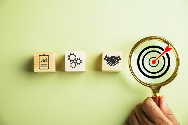 The photograph features a businessman using a magnifying glass to examine the target goal icon, underscoring the importance of strategic planning and global business leadership for success. - Foto, imagen