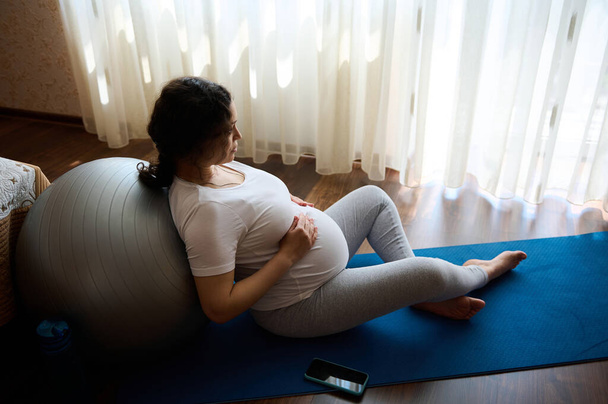 Authentic portrait of delightful pregnant woman, expectant mother holding her hands on her big belly, relaxing leaning on a fit ball after prenatal fitness at home, enjoying tranquil homely atmosphere - Photo, Image