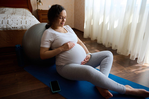 Grossesse en santé, maternité, concept de procréation. Portrait d'une future femme sportive aimante attentionnée attendant le nouveau-né, tenant ses mains sur son ventre, le caressant doucement, assise sur un tapis de yoga - Photo, image