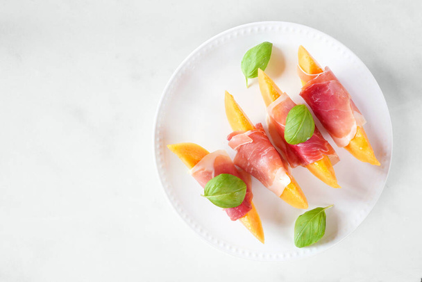 Fresh cantaloupe melon with prosciutto and basil. Overhead view on a white marble background. - Foto, imagen