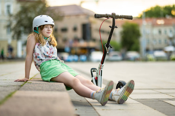 Adorable young girl riding her scooter in a city on sunny summer evening. Pretty preteen child riding a roller. Active leisure and outdoor sports for kids. - Foto, Bild