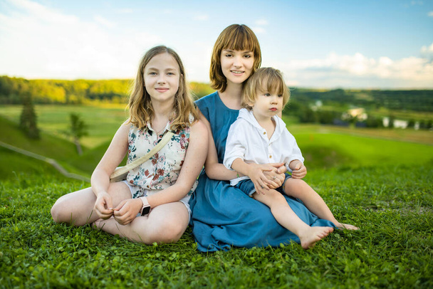 Two big sisters and their toddler brother having fun outdoors. Two young girls holding baby boy on summer day. Children with large age gap. Big age difference between siblings. Big family. - Photo, image
