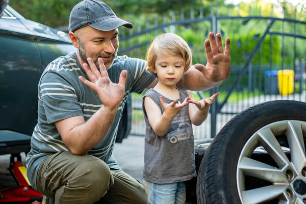 Cute toddler boy helping his father to change car wheels at their backyard. Father teaching his little son to use tools. Active parent of a small child. - Foto, Imagem