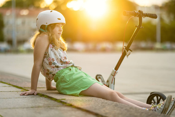 Adorable young girl riding her scooter in a city on sunny summer evening. Pretty preteen child riding a roller. Active leisure and outdoor sports for kids. - Foto, Imagen