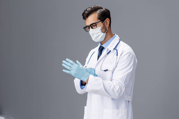 Médico masculino em um casaco branco e máscara médica e luvas estéreis, óculos olha para a câmera em um fundo isolado cinza, espaço de cópia, espaço para texto, saúde. Foto de alta qualidade - Foto, Imagem