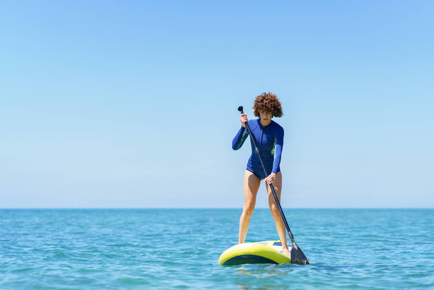 Opatrně žena surfařka v neoprénu stojící na SUP palubě a pádlování modré moře proti bezmračné modré obloze v slunečný letní den - Fotografie, Obrázek