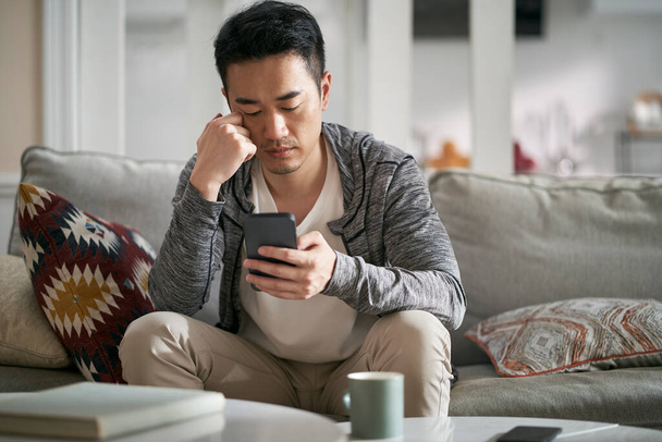 young asian man sitting on couch at home looking at cellphone looking serious - Photo, Image