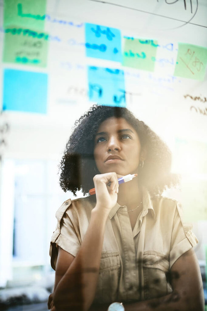 Gefangen im tiefen Ende ihrer Brainstorming-Sitzung. eine junge Geschäftsfrau beim Brainstorming mit Zetteln auf einer Glasscheibe in einem Büro - Foto, Bild