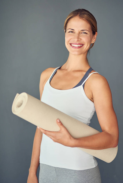 Vrolijke vrouw, yoga en portret glimlach met mat voor oefening, gezonde wellness of workout tegen een grijze studio achtergrond. Opgewonden vrouwelijke persoon of yogi lachend voor mindfulness, fitness of gezondheid van het lichaam. - Foto, afbeelding
