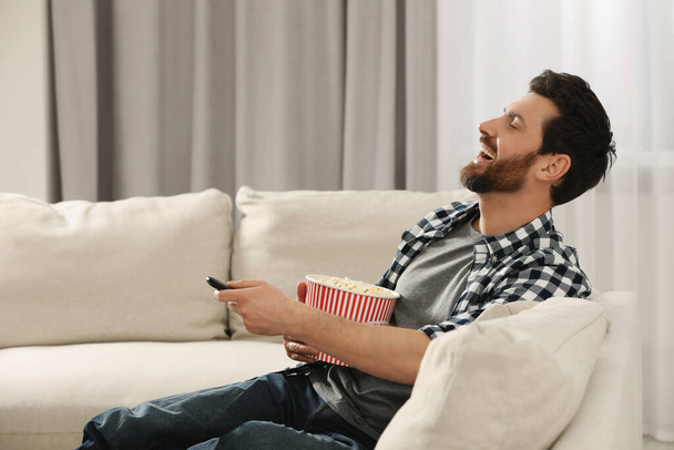 Laughing man watching TV with popcorn on sofa at home - Fotó, kép
