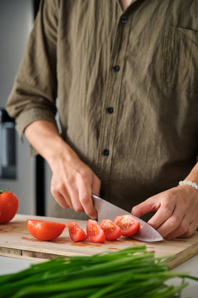 Αγνώριστος νεαρός που κόβει ντομάτες σε ένα ξύλο κοπής στην κουζίνα. - Φωτογραφία, εικόνα