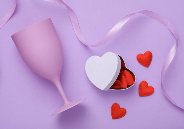 Anniversary, holiday concept. Plastic glasse and a box with many hearts and a satin ribbon on a lavender background. Valentine's Day. Top view - Photo, image