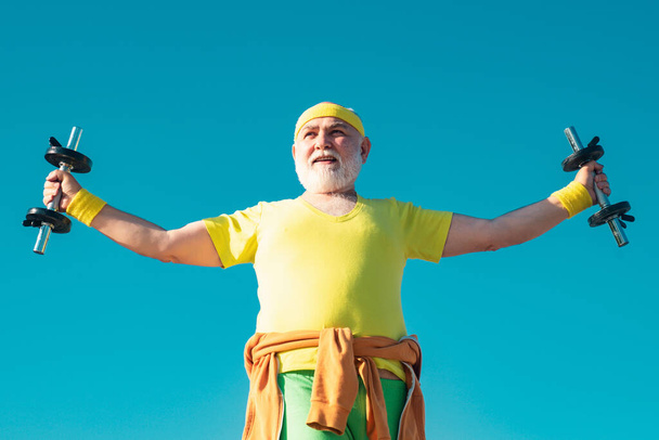 Homem mais velho no ginásio a treinar com pesos. Happy Senior man exercitando com haltere de elevação ao ar livre. Retrato de homem sênior segurando haltere
 - Foto, Imagem
