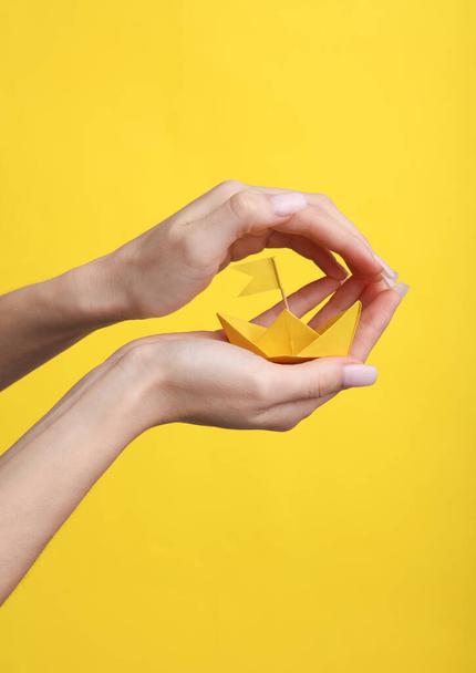 Hands holding and protects yellow paper boat on yellow background. Protection, insurance, security concept - Foto, afbeelding