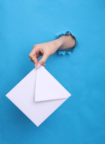 Female hand holds white envelope through torn hole in blue paper - Foto, Bild